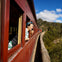 Two people lean out of windows of a red train traveling along a scenic route with trees and blue sky in the background.