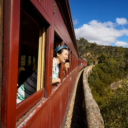Two people lean out of windows of a red train traveling along a scenic route with trees and blue sky in the background.
