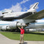A person in a red shirt stands in front of a large aircraft with a space shuttle mounted on top, displayed outdoors.