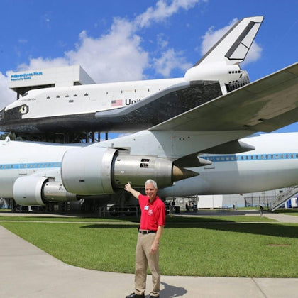 A person in a red shirt stands in front of a large aircraft with a space shuttle mounted on top, displayed outdoors.