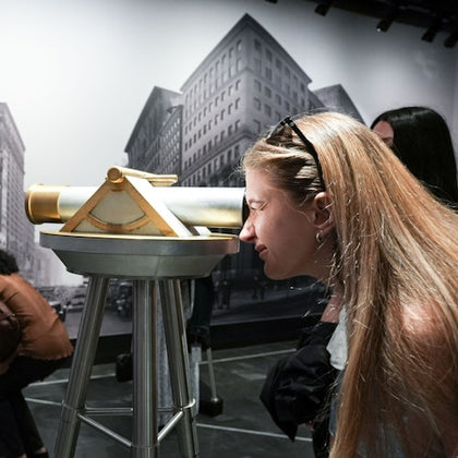 A woman looks through a mounted telescope in a museum with a black-and-white cityscape background.