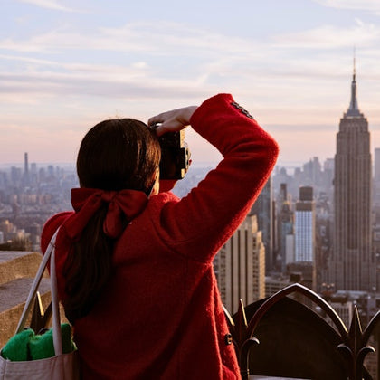 A person in a red coat takes a photo of a city skyline with the Empire State Building at sunset.
