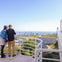 Two people stand on a terrace overlooking a cityscape, with railing and a clear blue sky in the background.