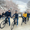 Three people on bikes, wearing helmets, pose on a path lined with blooming cherry blossoms. Other people walk in the background.