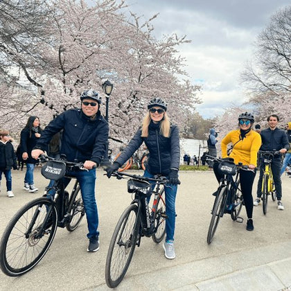 Three people on bikes, wearing helmets, pose on a path lined with blooming cherry blossoms. Other people walk in the background.