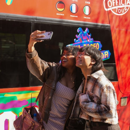 Two people smiling while taking a selfie in front of a colorful tour bus on a sunny day.