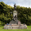Two people rest near a statue with the name "Garfield" on it. Two bicycles are leaned against the statue's base. Trees are in the background.