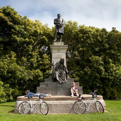 Two people rest near a statue with the name "Garfield" on it. Two bicycles are leaned against the statue's base. Trees are in the background.