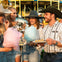 Group of people in casual attire enjoying cotton candy and food at an outdoor fairground with a carousel in the background.
