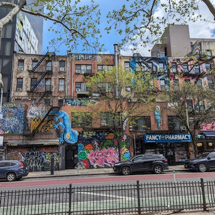 Street scene with brick buildings covered in colorful graffiti, a pharmacy storefront, and cars parked along the road.