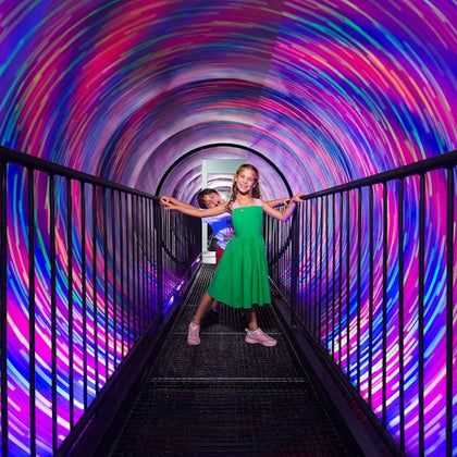 Two girls stand on a bridge inside a tunnel with swirling, colorful lights.