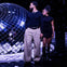Two individuals stand near a large reflective disco ball in a dark setting with reflective lighting effects.