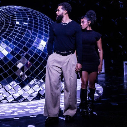Two individuals stand near a large reflective disco ball in a dark setting with reflective lighting effects.