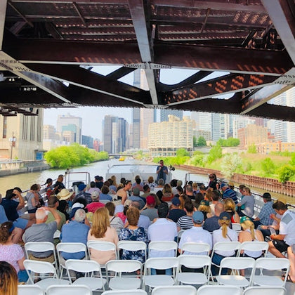 A group of people sitting on white chairs on a boat under a bridge, with a cityscape and waterway in the background.