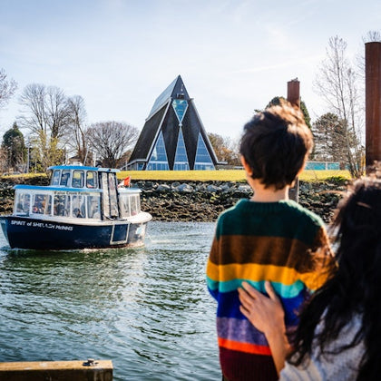 A person and a child stand by the water, watching a small boat. A modern triangular building is visible in the background.
