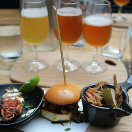 A meal with a small sandwich, meat dishes, and a flight of beer served on a wooden table.