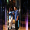 Two people, a woman and a man, stand together smiling and holding drinks in a reflective environment with colorful lighting.