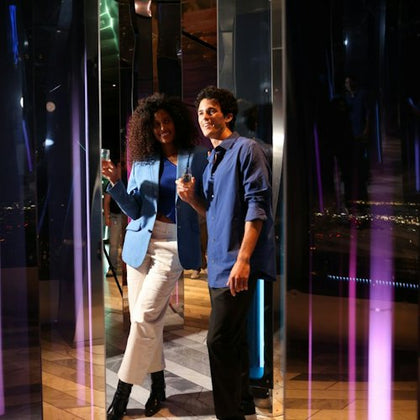 Two people, a woman and a man, stand together smiling and holding drinks in a reflective environment with colorful lighting.