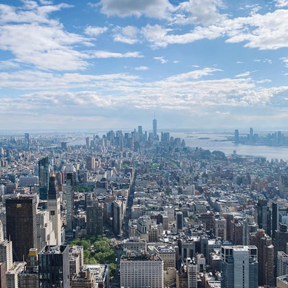 Aerial view of a cityscape featuring tall buildings, a distant skyline, and a river under a partly cloudy sky.