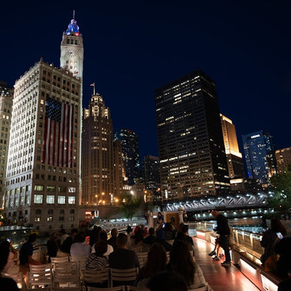 A group of people seated on a boat at night, with illuminated skyscrapers and a lit bridge in the background.