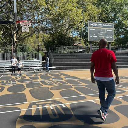A man in a red shirt walks on a patterned outdoor basketball court with three people and a scoreboard visible in the background.