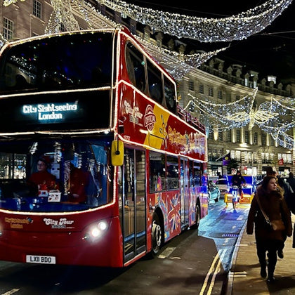 A red double-decker bus with "City Sightseeing London" displayed, decorated streets with lights, and pedestrians at night.