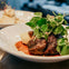 Plate of grilled meat topped with greens, served with mashed potatoes and sauce in the background on a dining table.