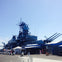 Large battleship docked at a pier with blue buildings and visitors walking nearby under a clear, sunny sky.