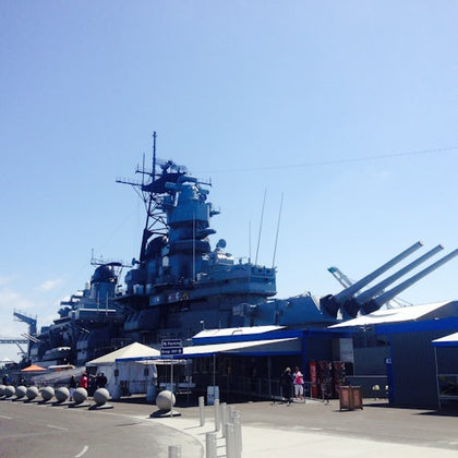 Large battleship docked at a pier with blue buildings and visitors walking nearby under a clear, sunny sky.