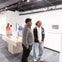 Two individuals stand in a museum, observing exhibits displayed in glass cases on a blue-carpeted floor.