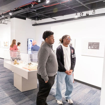 Two individuals stand in a museum, observing exhibits displayed in glass cases on a blue-carpeted floor.