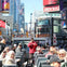 A group of people on an open-top tour bus in a city, with urban buildings and signs visible in the background.