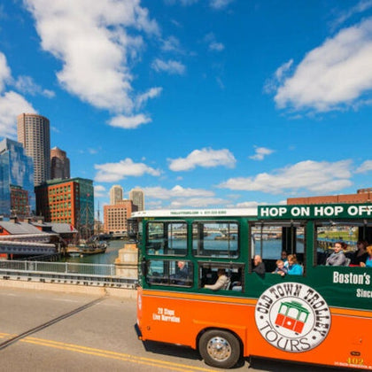 A vibrant orange and green trolley labeled "Old Town Trolley Tours" drives on a street with a city skyline and blue sky.