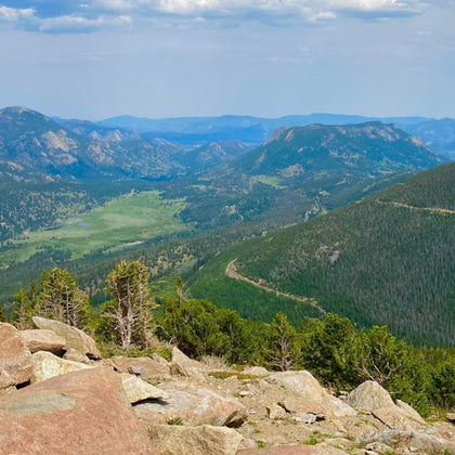 A scenic mountain landscape with rocky foreground, green forested slopes, and a valley stretching into the distance.