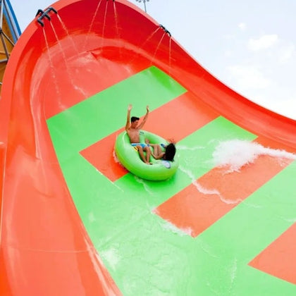 Two people ride a green inflatable tube down a brightly colored orange and green water slide with splashing water.
