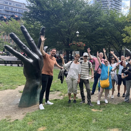 A group of people posing with a large hand sculpture in a park, with trees and buildings in the background.