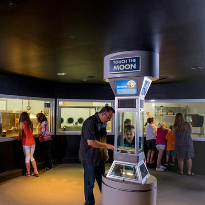 People in a museum exhibit interacting with a display labeled 'Touch the Moon,' with other exhibits visible in the background.