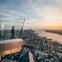 A person standing on a glass observation deck overlooking a cityscape and a river at sunset.