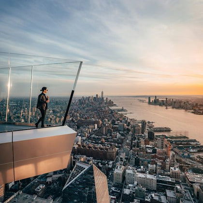 A person standing on a glass observation deck overlooking a cityscape and a river at sunset.