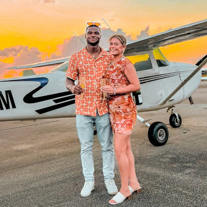 A couple in matching orange outfits stand in front of a small airplane during sunset, holding drinks and smiling.