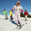 Four people skiing on a sunny snow-covered slope, wearing colorful ski outfits with a chairlift and mountains in the background.