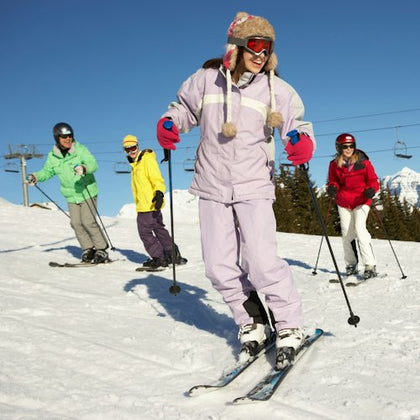Four people skiing on a sunny snow-covered slope, wearing colorful ski outfits with a chairlift and mountains in the background.