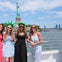 Four women posing on a boat in front of the Statue of Liberty with a clear sky and water in the background.