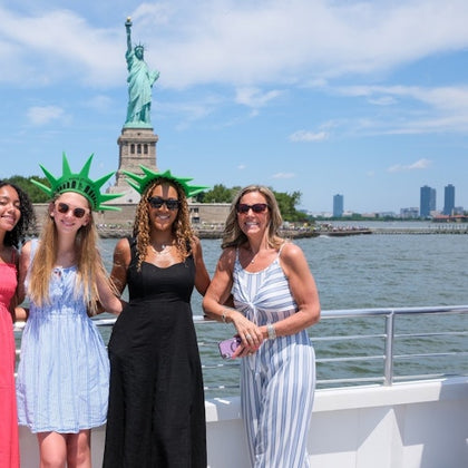Four women posing on a boat in front of the Statue of Liberty with a clear sky and water in the background.