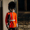 A guard in a red uniform and bearskin hat stands upright with a rifle, next to a stone wall and window.