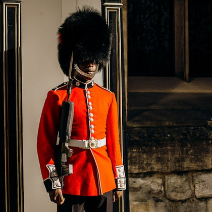 A guard in a red uniform and bearskin hat stands upright with a rifle, next to a stone wall and window.