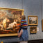 A child on an adult's shoulders viewing a large, ornate painting in an art gallery, with other framed artwork on the wall.