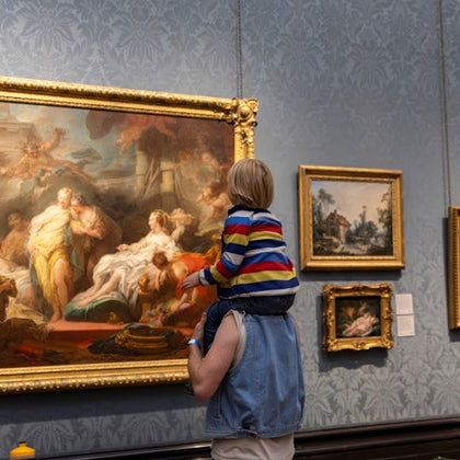 A child on an adult's shoulders viewing a large, ornate painting in an art gallery, with other framed artwork on the wall.