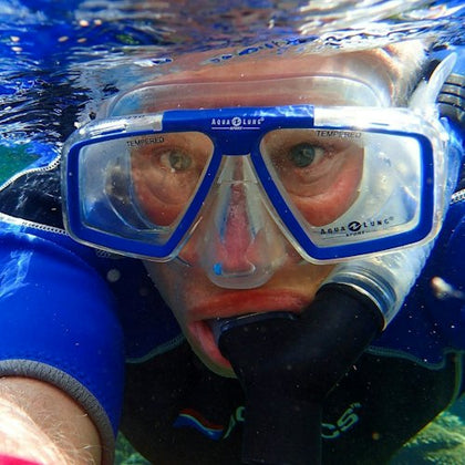Person wearing a blue wetsuit and blue diving mask with a snorkel, partially submerged in water, looking directly at the camera.