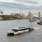 A modern ferry labeled "Uber Boat" travels on a river with Tower Bridge and city buildings in the background.